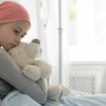 Weak girl with cancer wearing pink headscarf and hugging teddy bear